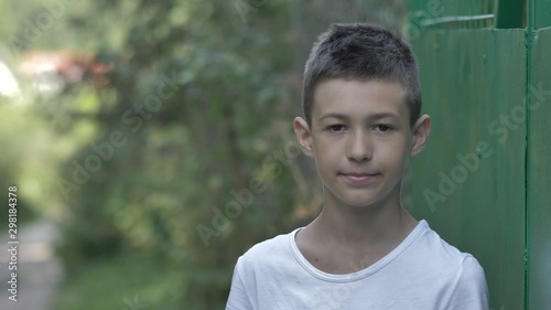 portrait of a funny boy outdoors near the fence stands and looks at the camera in the country photo