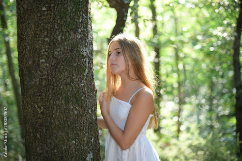 Girl leaning on a tree in a forest in a white dress and looking