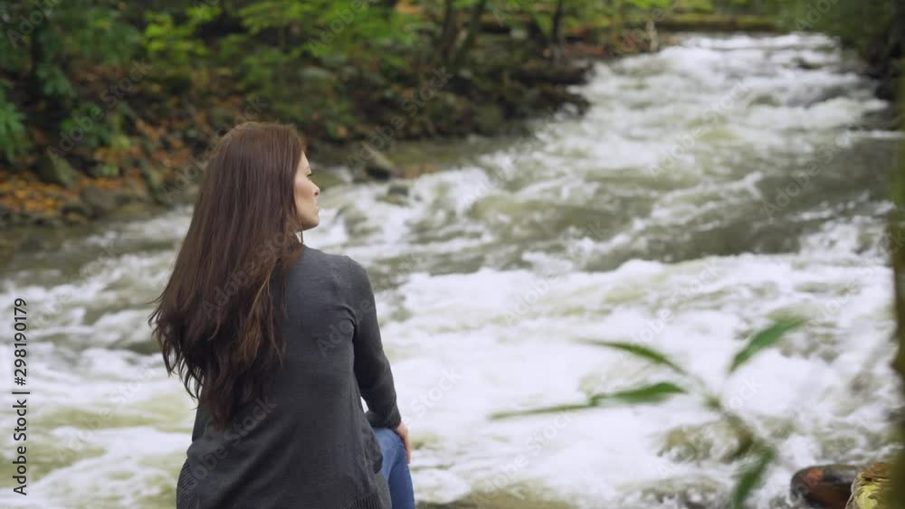 Young woman traveling alone by a flowing river with rapids contemplating and meditating  about peaceful thoughts while looking at the flowing water on an overcast day in the forest
