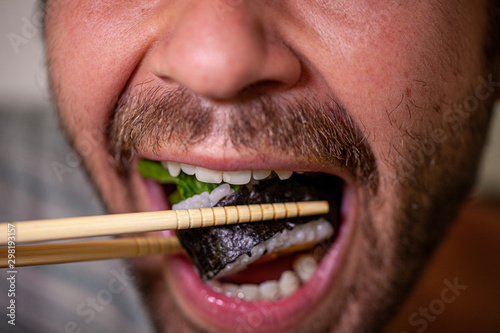 Close up of a mouth of a bearded caucasian man eating homemade sashimi sushi roll with chopsticks © Maarten Zeehandelaar