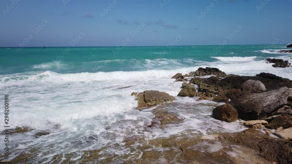 Turquoise water of the Caribbean crashing over rocks along and island shoreline. Bright blue sky and clear warm sea with white foam from waves breaking on a rocky beach. PAN RIGHT, slow motion. Turquoise water of the Caribbean crashing over rocks along and island shoreline. Bright blue sky and clear warm sea with white foam from waves breaking on a rocky beach. PAN RIGHT, slow motion.
