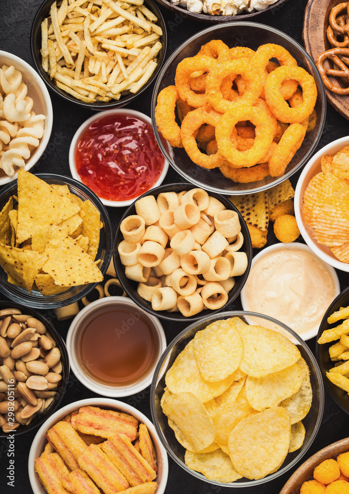 All classic potato snacks with peanuts, popcorn and onion rings and salted pretzels in bowl plates on black background. Twirls with sticks and potato chips and crisps with nachos and cheese balls.