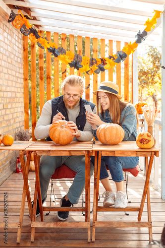 Halloween Preparaton Concept. Young couple sitting at table at porch making jack-o'-lantern man drawing face on pumpkin while woman taking photo on smartphone cheerful