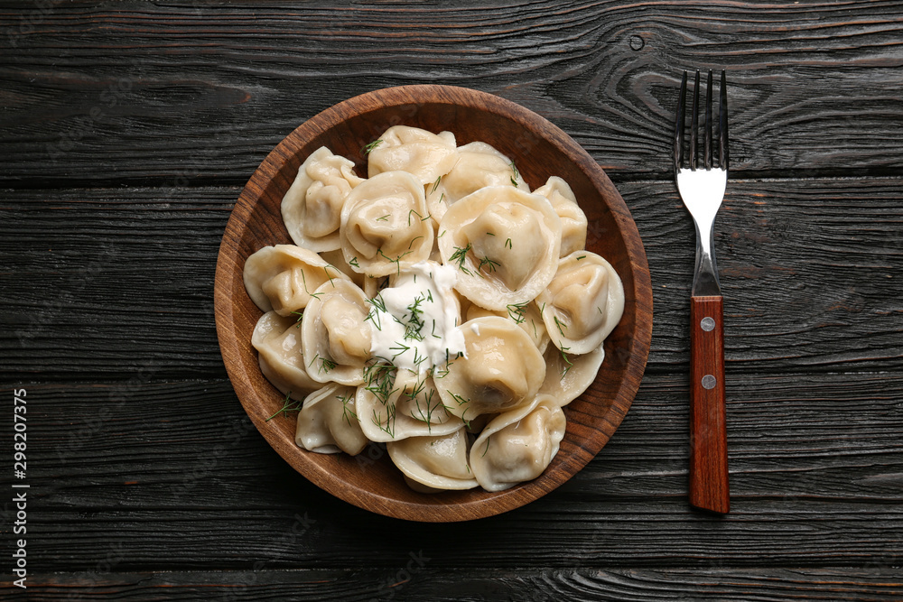 Tasty dumplings with sour cream in bowl on black wooden table, flat lay