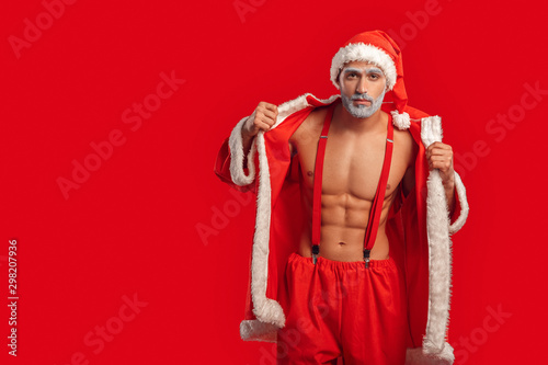 Christmas Freestyle. Young Santa Claus bare muscular upper body in hat standing isolated on red putting on coat pensive copy space