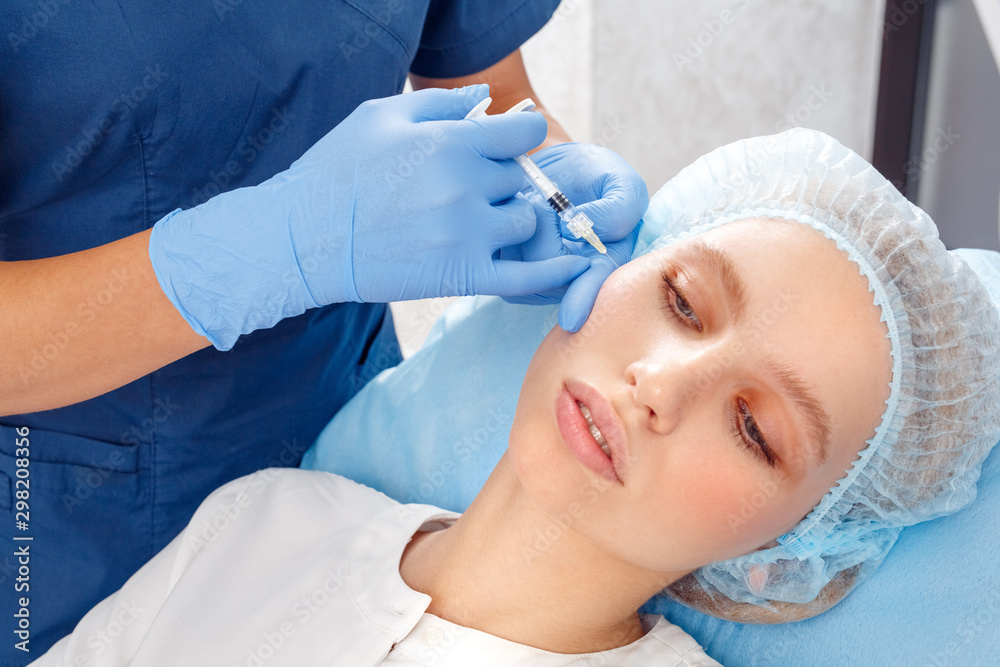 Cosmetology Service. Young woman at beauty clinic lying relaxed while doctor in gloves doing injection of collagen into cheek close-up