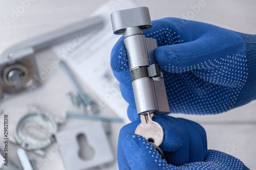 The guy holds in his hands in blue gloves the core of the lock and door keys on a light background. Repair in the house. Installing a new lock on the door.