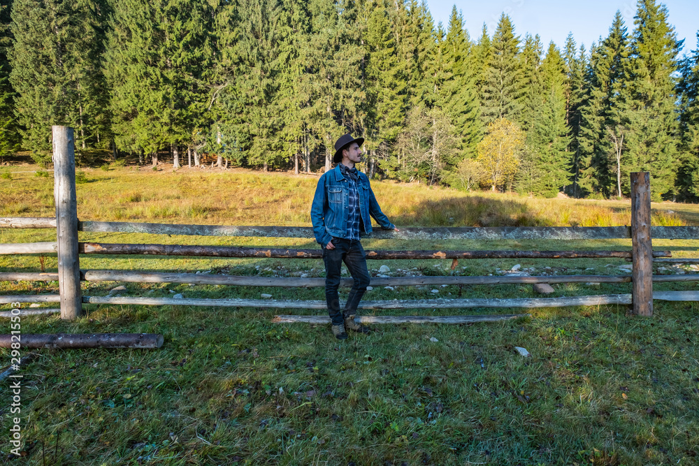 Young male farmer in a denim jacket and cowboy hat stand near the wooden  fence against forest 