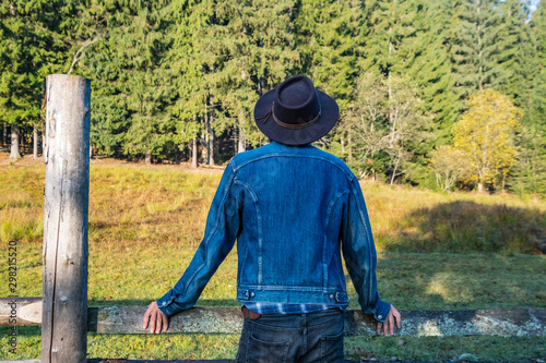 Young male farmer in a denim jacket and cowboy hat stand near the wooden fence against forest 
