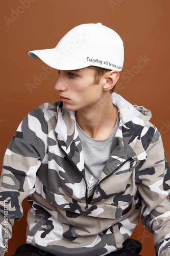 Profile photo of European man with short fair hair in a camouflage hoodie, a gray tee-shirt and white baseball cap with black lettering. The man is posing on the brown background. photo