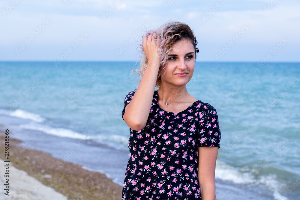 beautiful female with curly hairs in black dress in flowers is standing near water on the beach. Orange sunset light on the face beautiful female with curly hairs in black dress in flowers is standing near water on the beach. Orange sunset light on the face