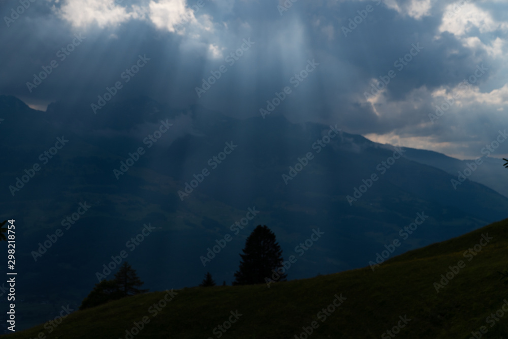 Sun rays shining trough the clouds in the mountains, a hill and trees in front
