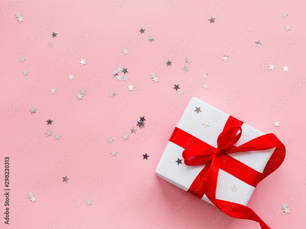 Present in white wrapping paper on pink backdrop with silver star confetti. Holiday gift with red ribbon and bow. Top view, flat lay.