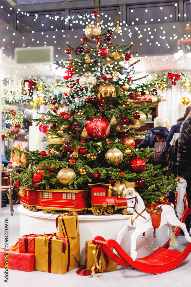 Christmas tree with colorful decorative balls. New Year decorations and gifts - rocking horse, presents ni red and gold wrapping paper. Festive interior of a shop.