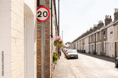 Terraced street showing a 20mph speed limit sign. The historic, old houses are seen together with parked vehicles. often used as a busy commuter road.