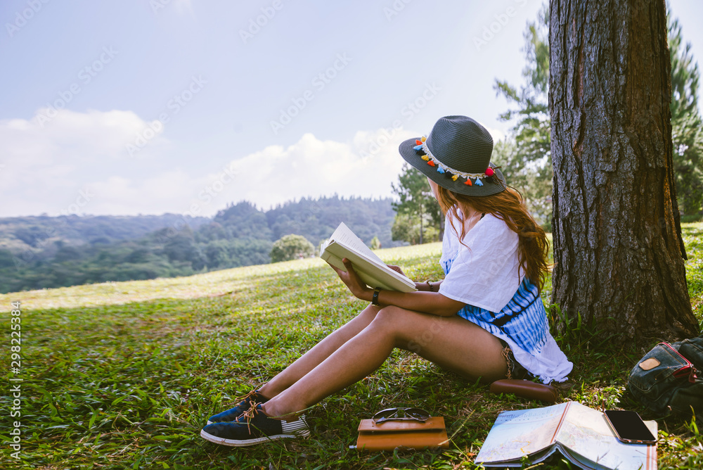 Fototapeta premium Asian woman travel nature. Travel relax. Girl sitting reading a book Under the tree. Beautiful girl in autumn forest reading a book Nature Education and Write a note.