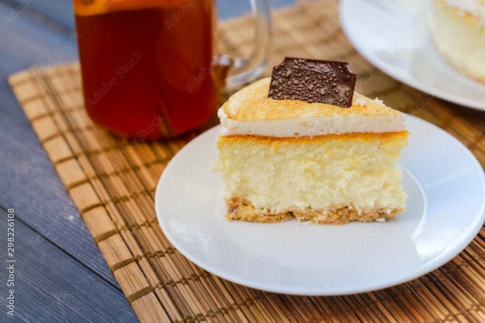 Classic cheesecake cake on a dish, a piece of cake on a saucer, a spoon, tea in a mug, everything on a straw napkin on a dark wood tabletop Classic cheesecake cake on a dish, a piece of cake on a saucer, a spoon, tea in a mug, everything on a straw napkin on a dark wood tabletop