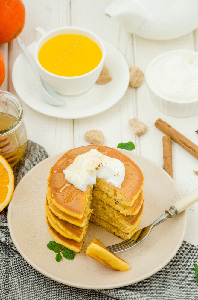Stack of spicy pumpkin pancakes on a plate with honey and whipped cream on a white wooden background.  Rustic style. Fragrant autumn breakfast. Breakfast for Halloween.
