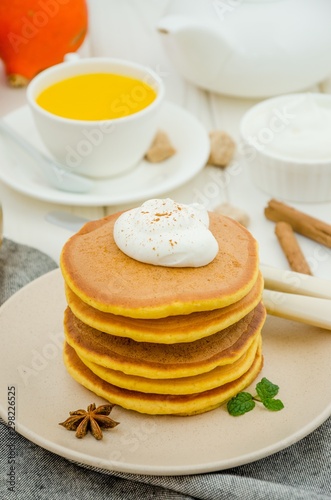 Stack of spicy pumpkin pancakes on a plate with honey and whipped cream on a white wooden background.  Rustic style. Fragrant autumn breakfast. Breakfast for Halloween.