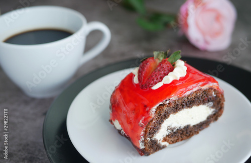 chocolate cake with strawberry jam sauce topped with whipped cream and fresh strawberry chopped served on white plate with a cup of black coffee  blur rose at background
