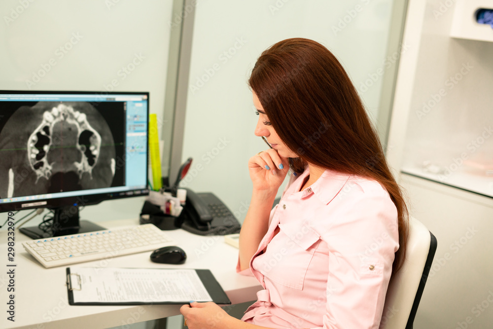 A female dentist doctor is sitting at a table, on a computer a CT scan of the jaw. The doctor is dressed in professional clothes. A female dentist doctor is sitting at a table, on a computer a CT scan of the jaw. The doctor is dressed in professional clothes.