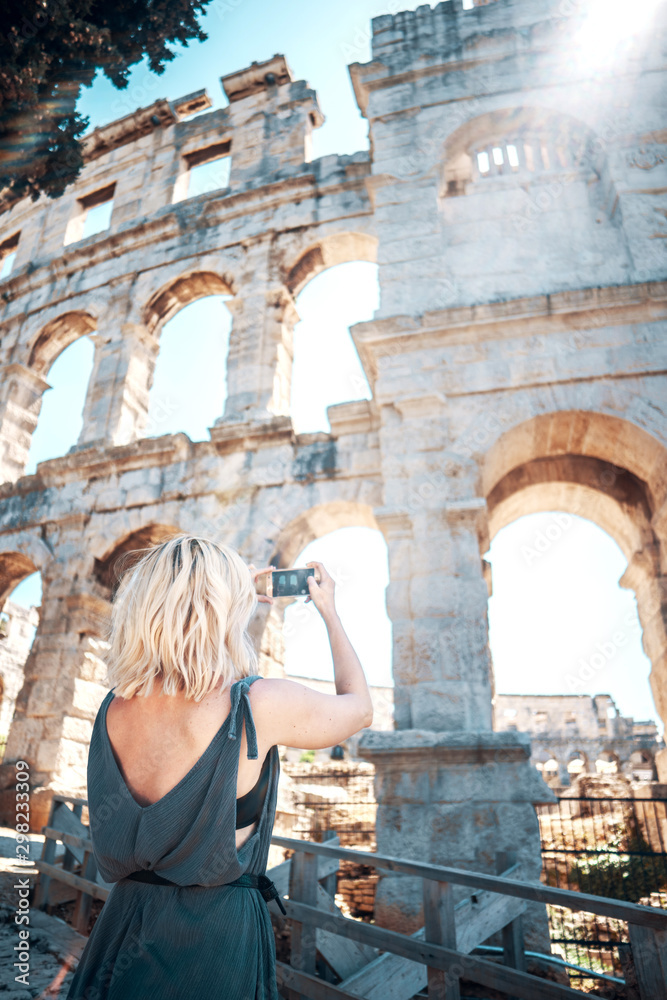 Young female tourist takes pictures of Pula Arena, Croatia with her smartphone on beautiful sunny day. Shot from behind. European travel destinations concept