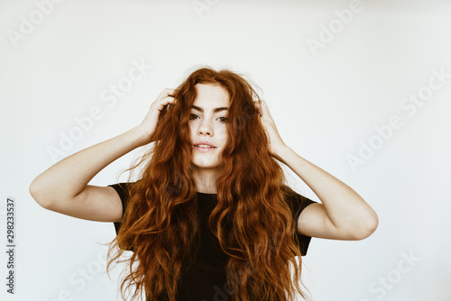 Lovely young girl with long curly red hair with freckles on her face in a black T-shirt on a white background holds her hands by her hair and poses