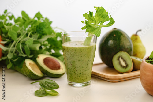 Green smoothie with a stalk of celery in a glass cup. On the table is an avocado.