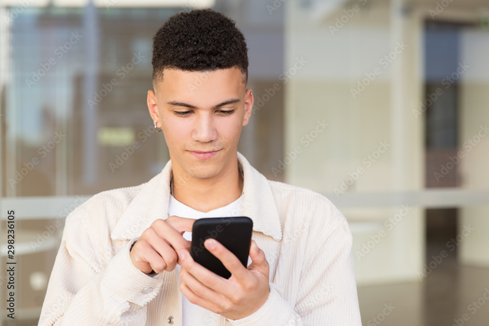 Handsome young man using smartphone outdoor. Smiling young man with piercing standing on street and messaging via cell phone. Communication concept
