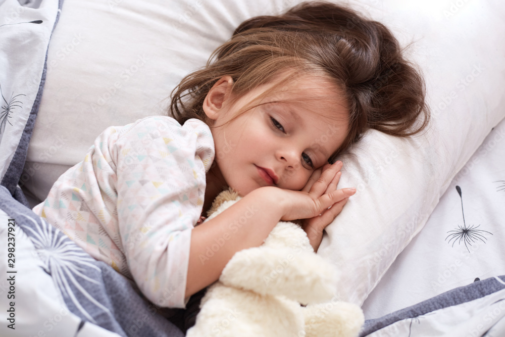 Little girl with white soft dog relaxing in bed, hugging her toy, keeps eyes opened lying on pillow under warm blanket, charming female kid with dark hair wearing white pajama. Morning time concept. Little girl with white soft dog relaxing in bed, hugging her toy, keeps eyes opened lying on pillow under warm blanket, charming female kid with dark hair wearing white pajama. Morning time concept.
