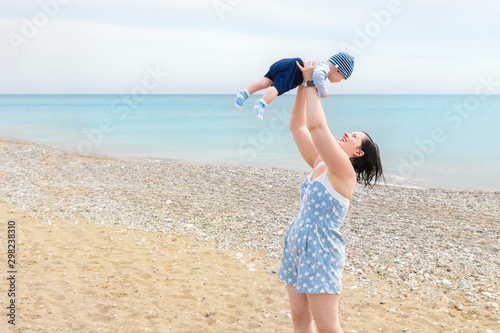 Young laughing mom holding her baby boy son on the beach at the sea vacation both wearing blue clothes