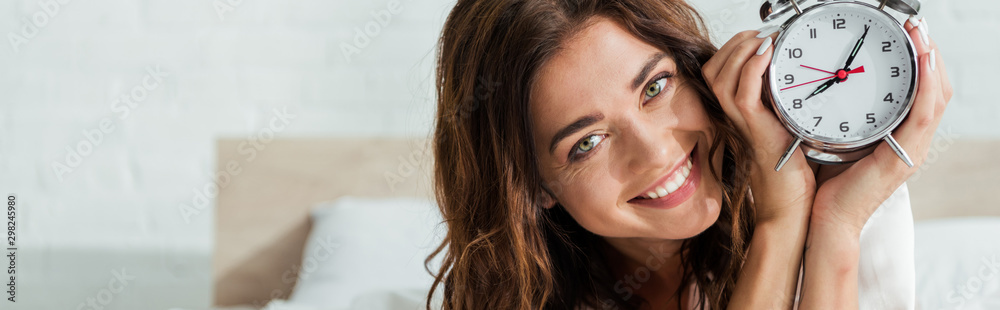 panoramic shot of attractive woman smiling and holding alarm clock at morning - obrazy, fototapety, plakaty 