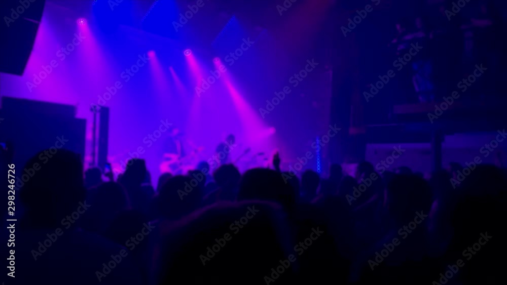 People dance and jump at a rock concert in a club, a band plays on stage, the background is colorful and blurred People dance and jump at a rock concert in a club, a band plays on stage, the background is colorful and blurred