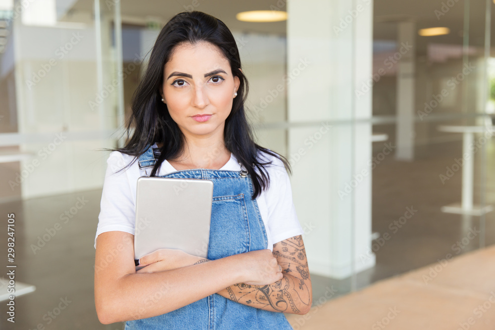 Serious woman with digital tablet looking at camera. Portrait of beautiful young brunette woman in denim overall holding tablet pc and looking at camera. Wireless technology concept Serious woman with digital tablet looking at camera. Portrait of beautiful young brunette woman in denim overall holding tablet pc and looking at camera. Wireless technology concept