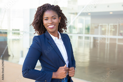 Happy confident professional posing near office building. Young African American business woman standing outside, adjusting formal jacket, looking at camera, smiling. Successful businesswoman concept