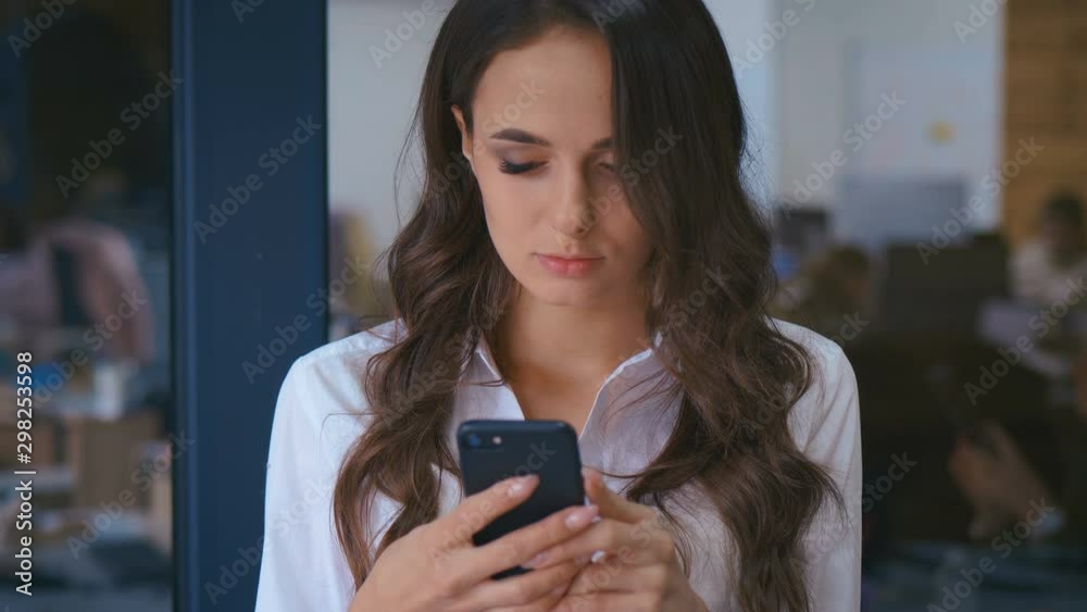 closeup of beautiful young business woman with smartphone texting messenger inside office in the background