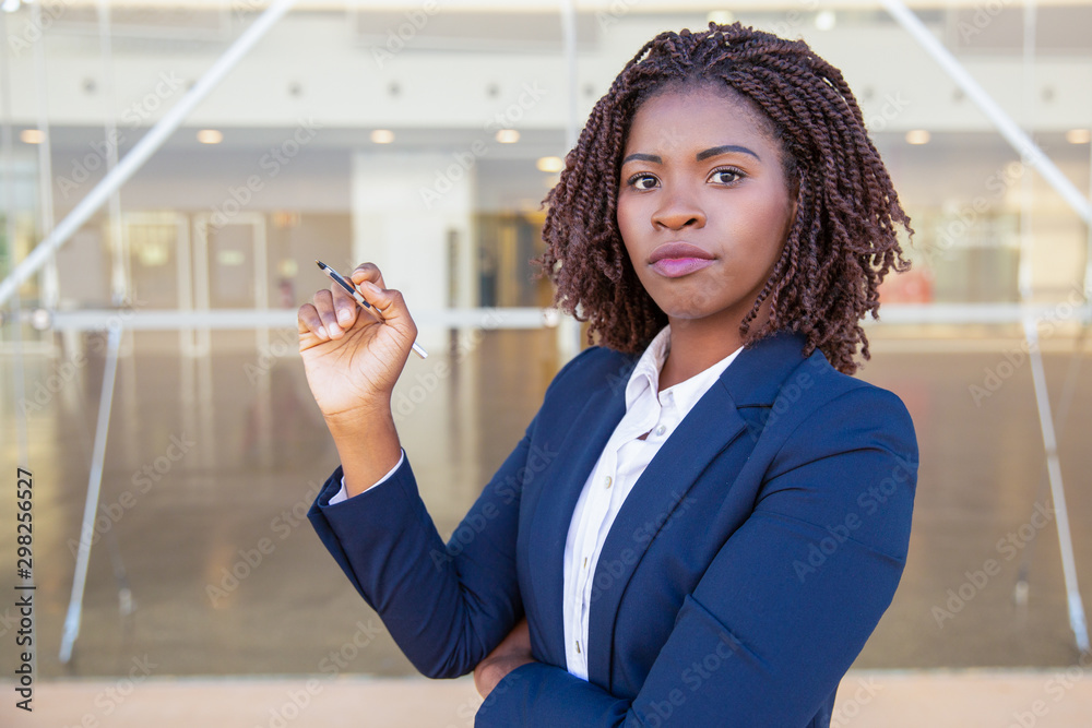Serious confident manager pointing at copy space. Young African American business woman holding pen, writing, looking at camera. Advertising concept Serious confident manager pointing at copy space. Young African American business woman holding pen, writing, looking at camera. Advertising concept