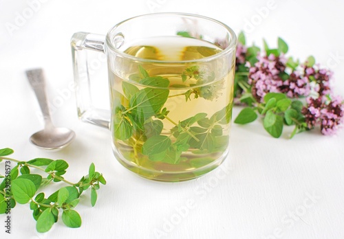 Fresh oregano tea in glass cup on white table, fresh herbs, spoon, natural home apothecary.