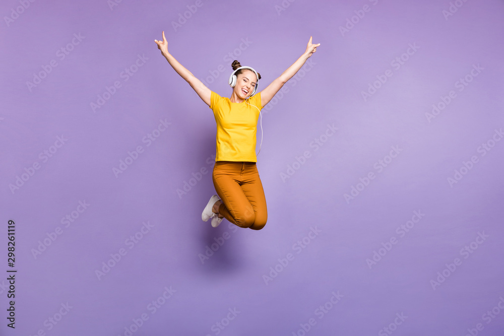 Full loength photo of joyful lady jumping high listening modern youngster playlist music through cool earflaps wear yellow t-shirt trousers isolated pastel purple background