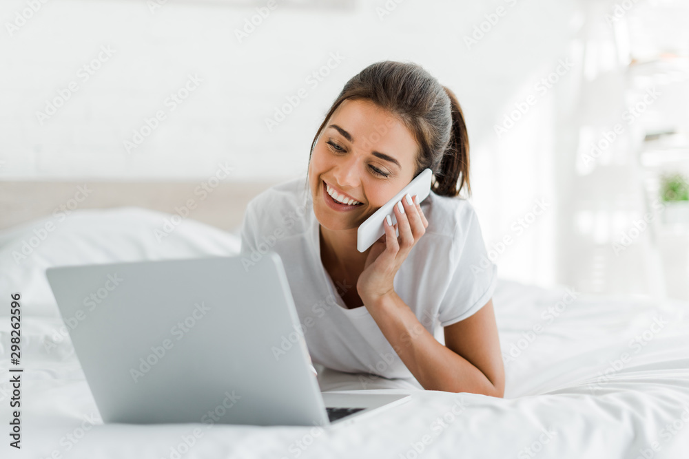 attractive smiling girl talking on smartphone and using laptop in bed in the morning attractive smiling girl talking on smartphone and using laptop in bed in the morning