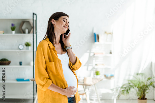 happy pregnant freelancer in yellow shirt talking on smartphone in home office