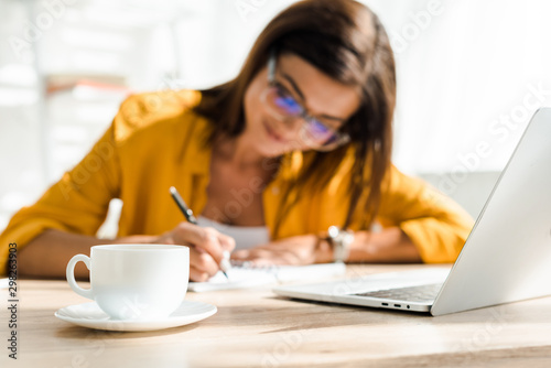 фотографія selective focus of coffee cup and beautiful freelancer writing in home office