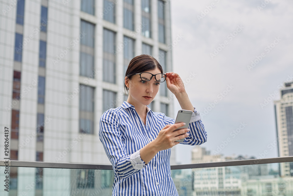Intelligent business woman with brown hair in glasses in a blue and white striped shirt with white cuffs on her sleeves looking surprise gray phone in her hand against the background of the city Intelligent business woman with brown hair in glasses in a blue and white striped shirt with white cuffs on her sleeves looking surprise gray phone in her hand against the background of the city