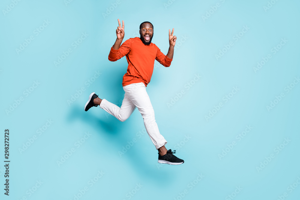 Full length body size photo of cheerful positive casual man running jumping mixed-race wearing white pants trousers sneakers showing double v-sign isolated vivid blue color background