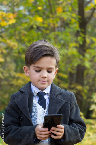 Kids and gadgets concept. A smiling happy schoolboy is standing in the street in a beautiful business suit holding a smartphone in his hands. A tie, a white shirt and autumn coat on him. Front view