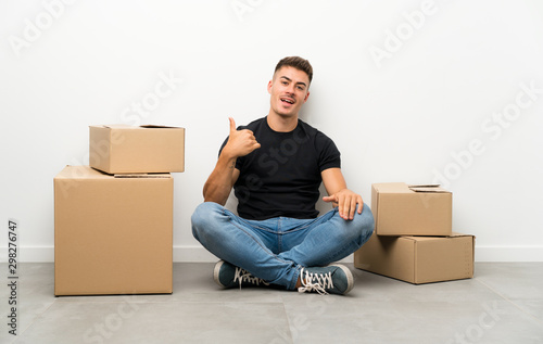 Handsome young man moving in new home among boxes making phone gesture