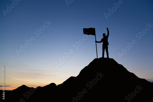 Silhouette of businessman holding a flag on top mountain, sky and sun light background. Business success and goal concept.