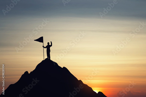 Silhouette of businessman holding a flag on top mountain, sky and sun light background. Business success and goal concept.