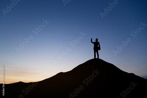 Silhouette of businessman raising his hand to celebrate success on top mountain, sky and sun light background.