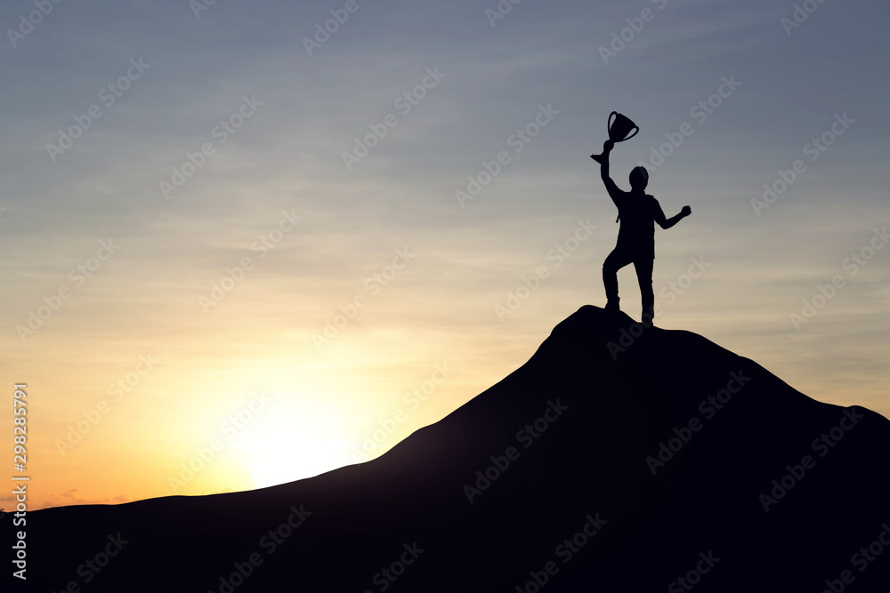 Silhouette of businessman with a championship cup celebrate success on top mountain, sky and sun light background.